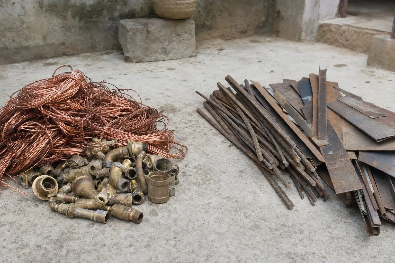 Sorted household scrap on a courtyard floor: copper wire and brass fittings in one pile, rusty iron rods and steel offcuts in another